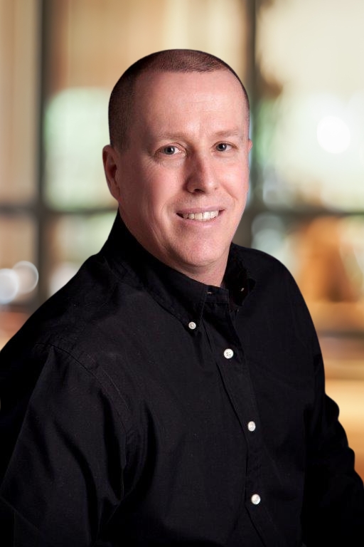 A man with short brown hair wearing a black button-up shirt smiles while facing the camera, with a softly blurred indoor background behind him.